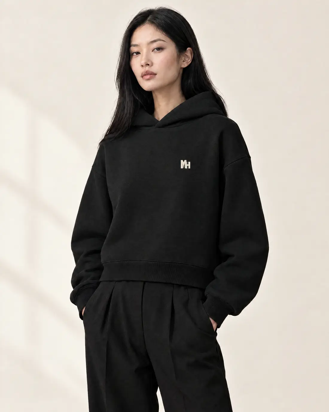 A woman with long black hair in a cropped black Melanated Humans hoodie with a small cream MH monogram at the left chest