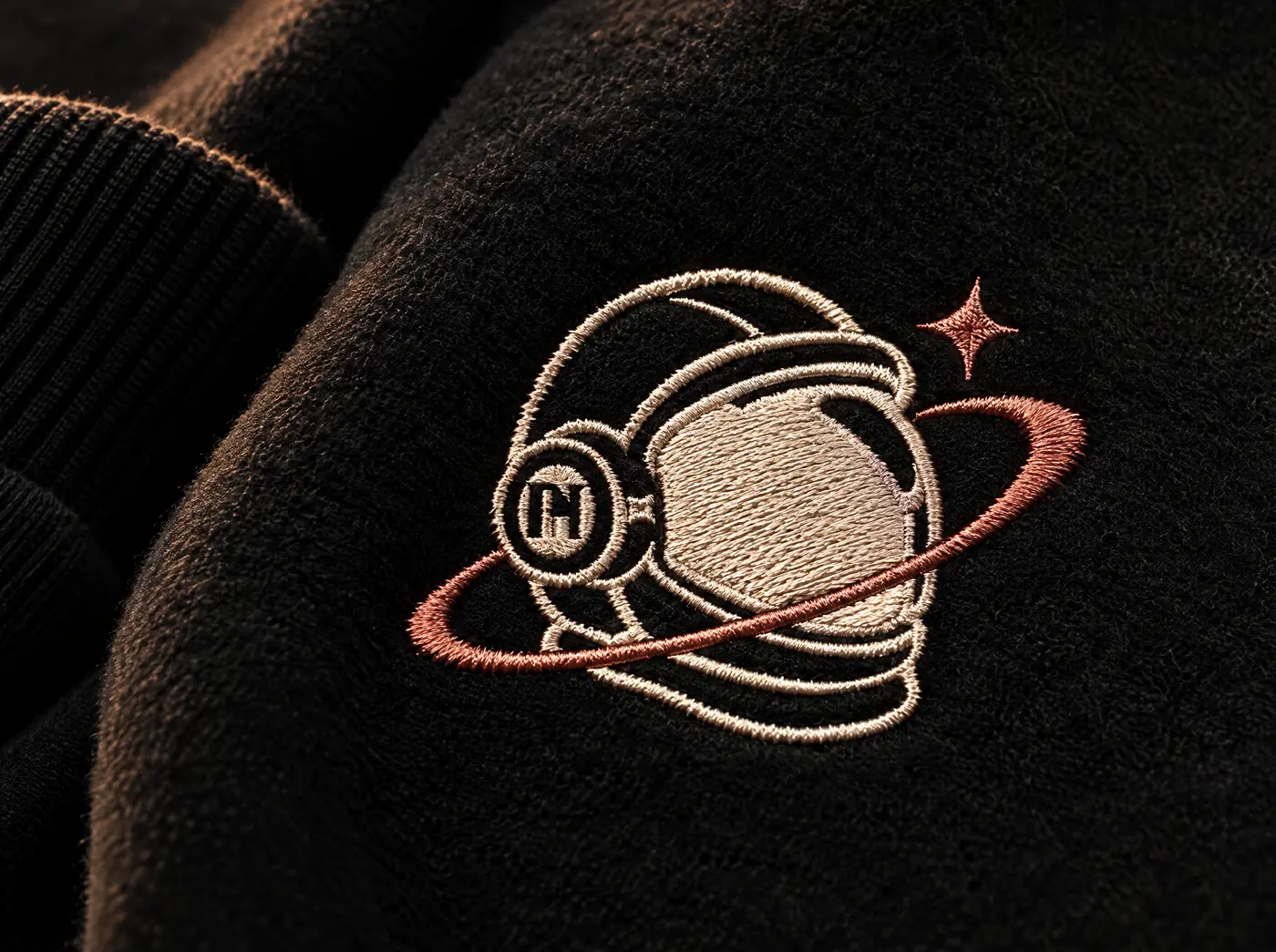 Embroidered hoodies close-up: the MH astronaut emblem stitched in cream and copper thread on black fleece